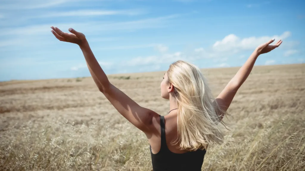 Ragazza bionda di spalle che alza le mani al cielo in un campo di avena, diventare liberi grazie alla pratica delle Costellazioni Familiari